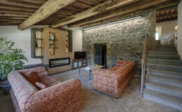 Cozy living room featuring two patterned sofas, a modern fireplace, and a staircase, complemented by natural wood beams and a stone accent wall. Perfect for relaxation and inviting gatherings.