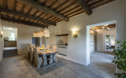 Modern dining area featuring a wooden table with eight chairs, rustic ceiling beams, and pendant lighting, leading to a well-equipped kitchen and a cozy living space.