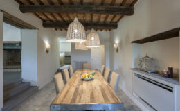 Spacious dining room featuring a rustic wooden table surrounded by upholstered chairs, illuminated by stylish pendant lights and complemented by a warm color palette and exposed wooden beams.
