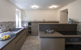 Modern kitchen interior featuring sleek cabinetry, an island with a cooktop, and a stylish backsplash. The space is bright and airy with natural light coming through a window, complemented by decorative items and fresh fruit on the counter. Ideal for contemporary home design inspiration.