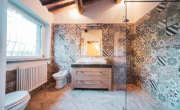 Modern bathroom interior featuring a decorative tiled wall, wooden vanity with sink, mirror, and shower area, complemented by natural light from a window.