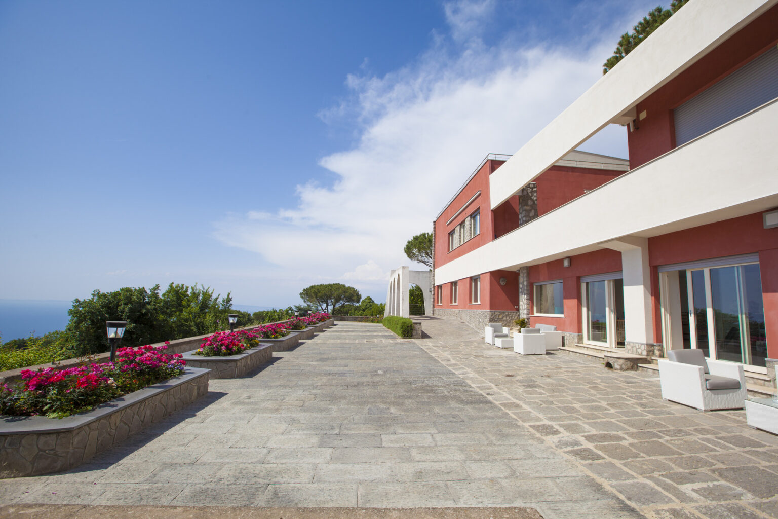 Modern villa exterior with a stone pathway lined with vibrant flowers, featuring large windows and a panoramic view of the ocean under a clear blue sky. | Only on SopranoVillas