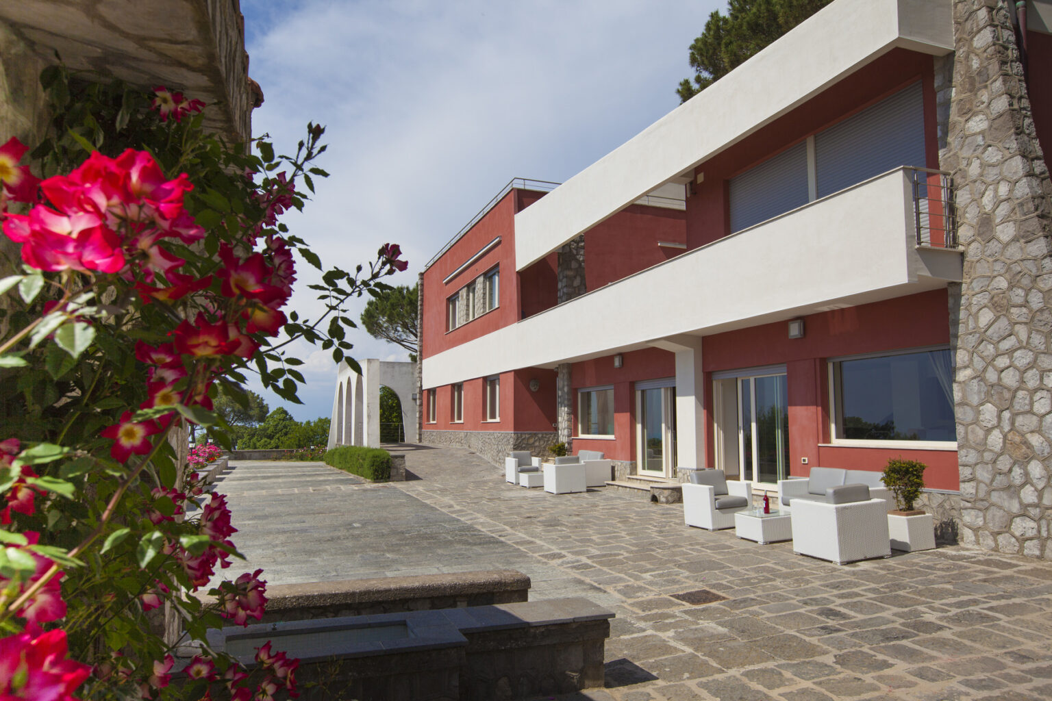 Modern villa with a stone and red exterior, featuring a spacious terrace adorned with white furniture and vibrant pink flowers in the foreground. | Only on SopranoVillas