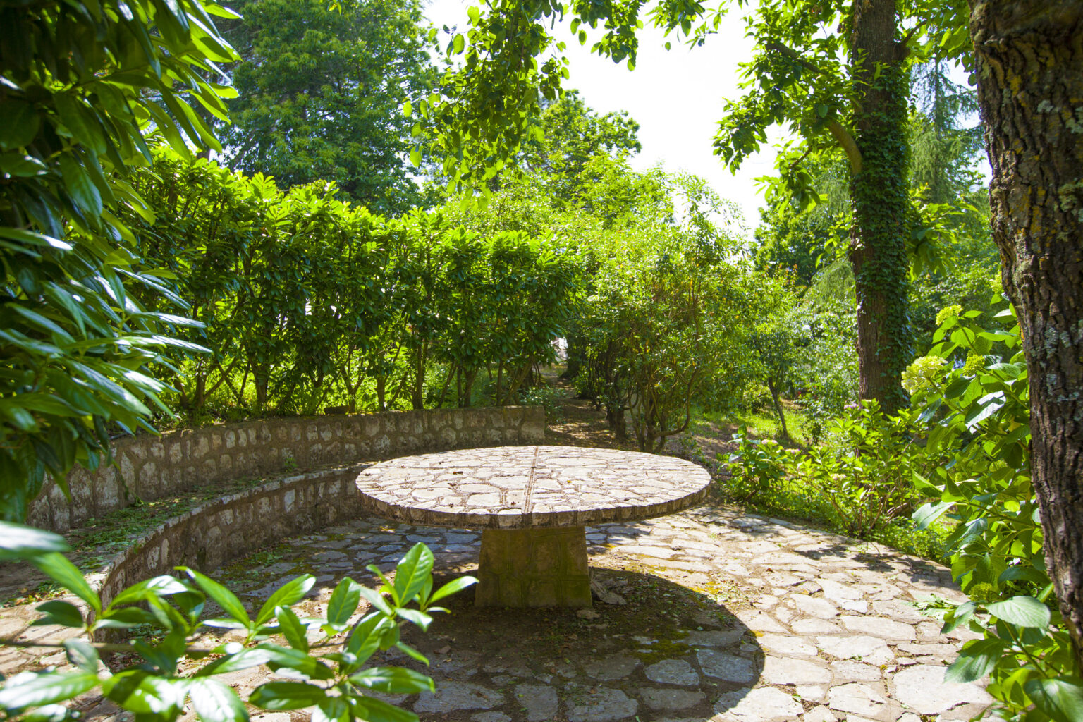 Serene garden scene featuring a stone table surrounded by lush greenery and trees, ideal for outdoor gatherings and relaxation. | Only on SopranoVillas