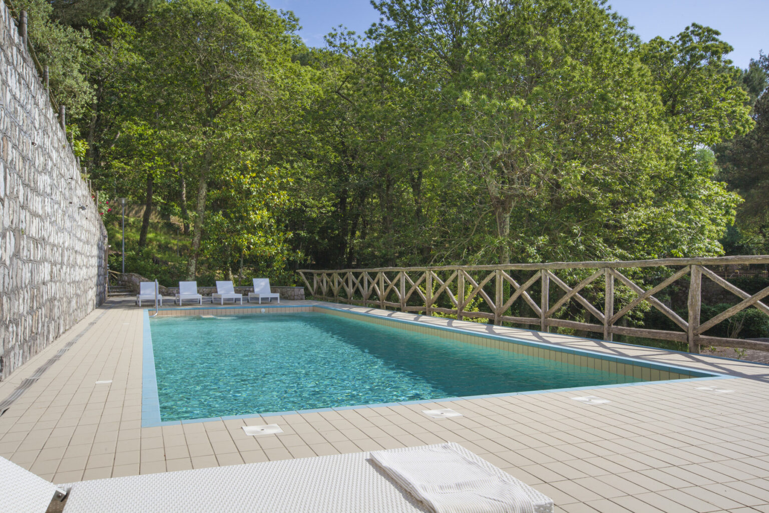 Serene outdoor swimming pool surrounded by lush greenery, featuring a stone wall and wooden railing, with white lounge chairs positioned for relaxation. Ideal for leisure and summer retreats. | Only on SopranoVillas