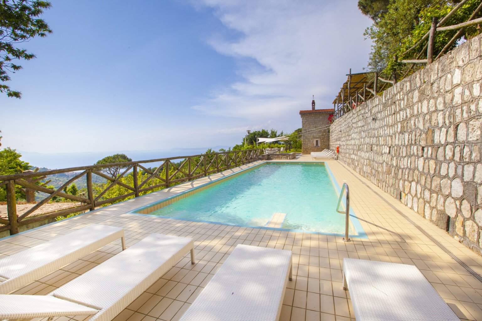 Scenic outdoor swimming pool with lounge chairs overlooking a lush landscape and distant ocean views, surrounded by stone walls and greenery. Ideal for relaxation and leisure in a tranquil setting. | Only on SopranoVillas