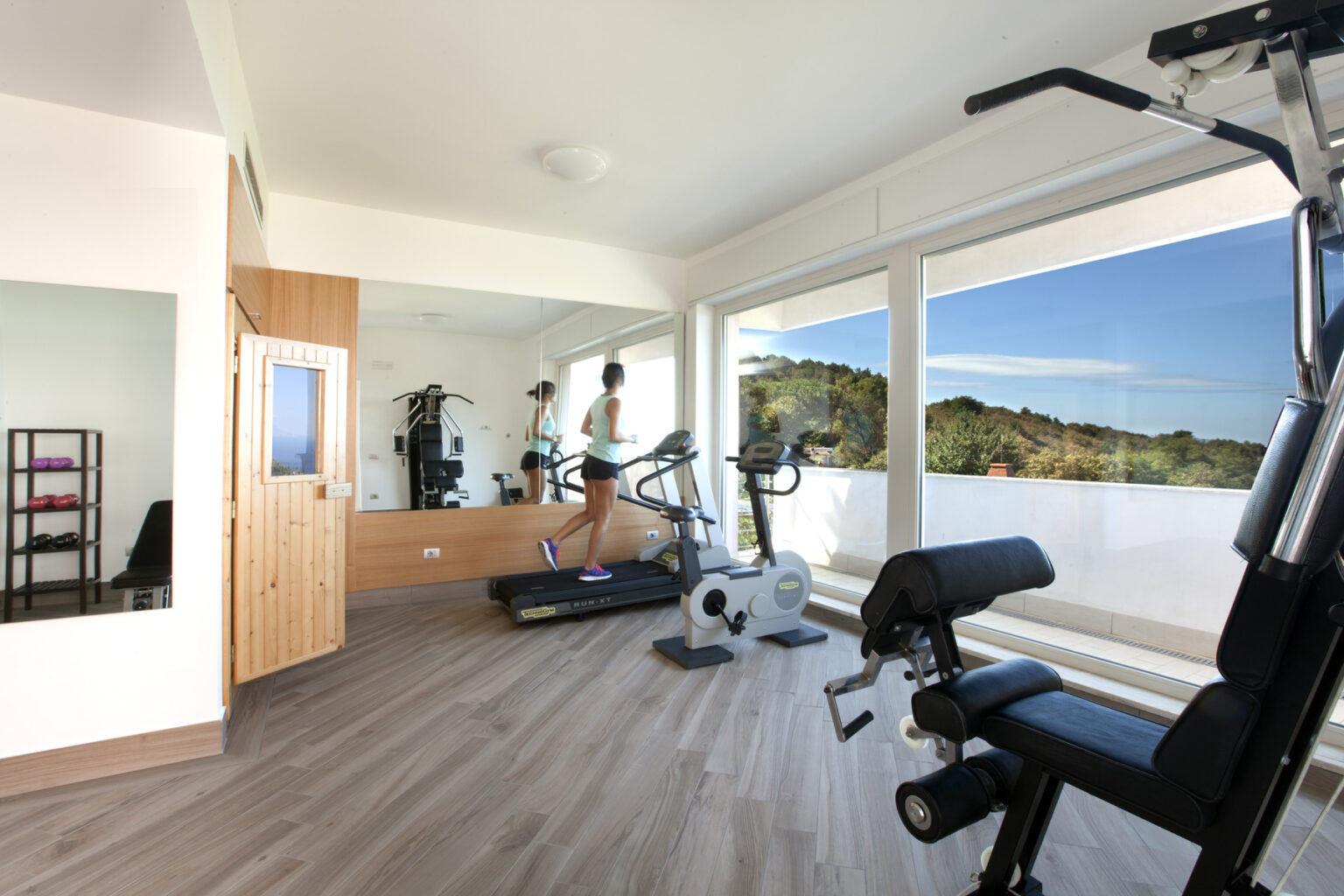 Bright and modern home gym with a treadmill and multi-gym equipment, featuring large windows overlooking a scenic landscape. A woman jogs on the treadmill, emphasizing an active lifestyle in a well-lit space. | Only on SopranoVillas