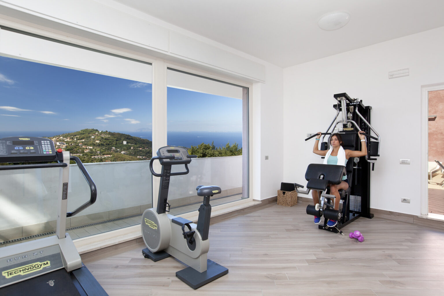 Home gym with panoramic ocean view featuring a treadmill, stationary bike, and multi-gym equipment. A woman exercises on the multi-gym, promoting fitness and wellness in a bright, modern space. | Only on SopranoVillas