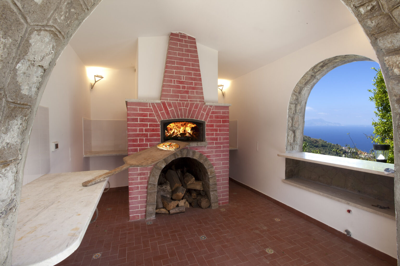 Rustic outdoor pizza oven made of red brick, featuring a wooden pizza peel with a freshly baked pizza, set in a modern kitchen with stone arches and a scenic ocean view through an arched window. | Only on SopranoVillas