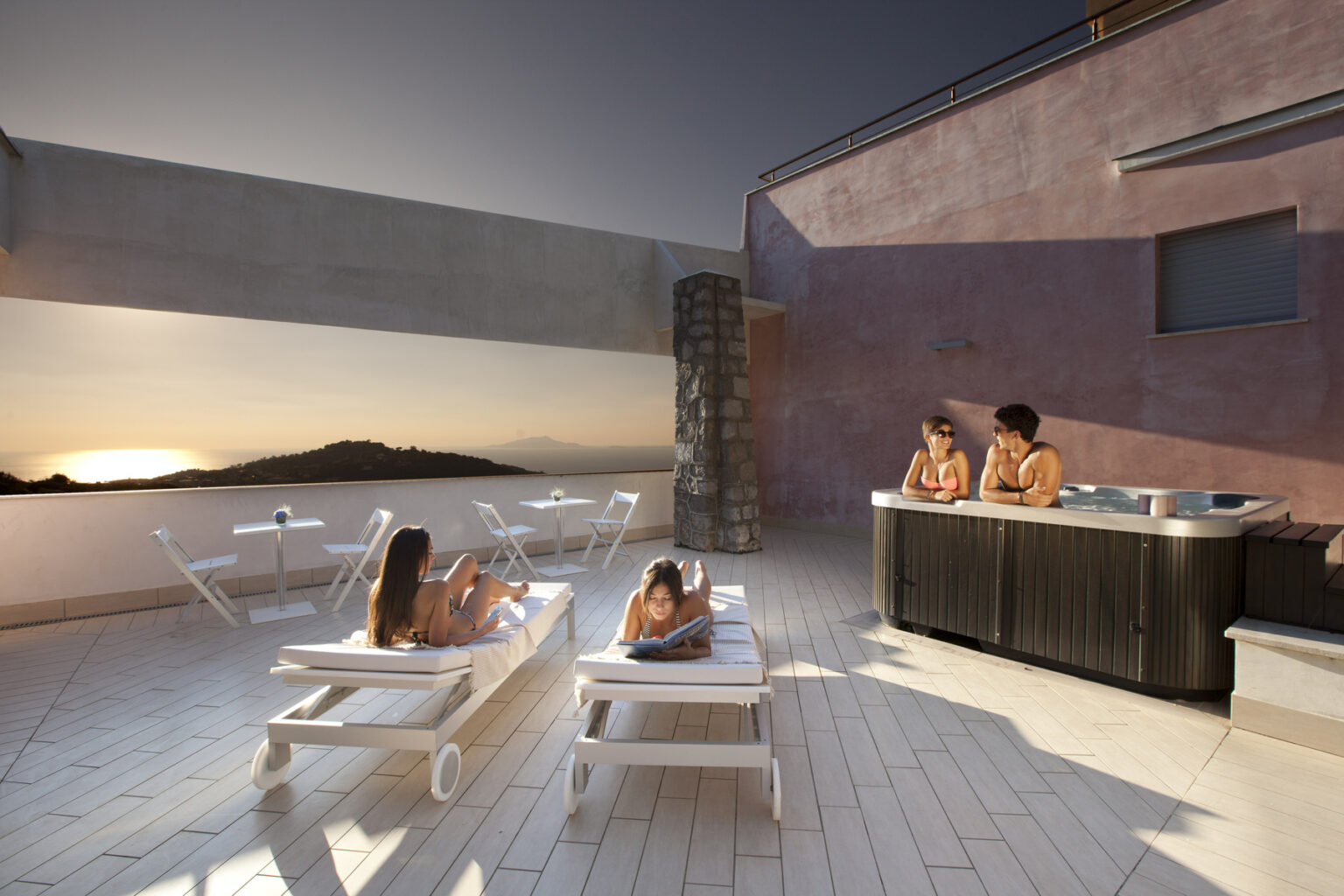 Two women relax in a hot tub on a terrace with a sunset view, while another woman lounges on a sunbed. The setting features modern architecture and a serene atmosphere, ideal for a luxurious getaway. | Only on SopranoVillas