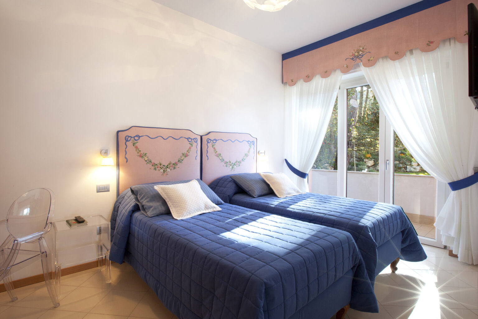 Bright and airy bedroom featuring two twin beds with blue bedding, decorative headboards, and large windows draped with sheer curtains, allowing natural light to fill the space. | Only on SopranoVillas