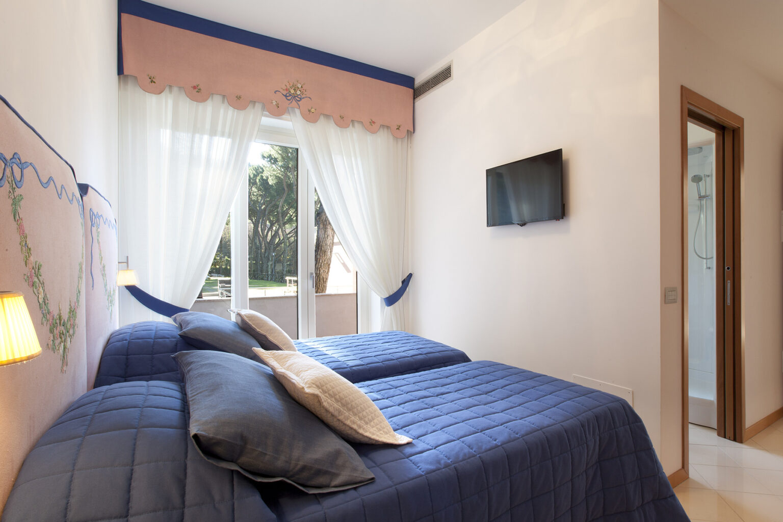 Cozy bedroom featuring two twin beds with blue bedding, a flat-screen TV mounted on the wall, and large windows with sheer curtains allowing natural light. The room includes a modern decor and a view of the outdoors. | Only on SopranoVillas