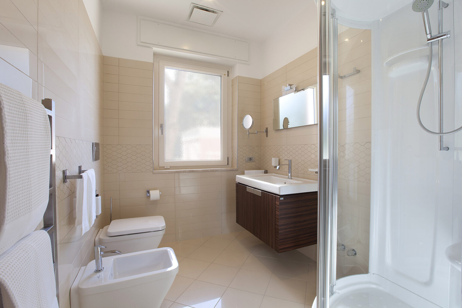 Modern bathroom featuring a glass shower enclosure, wall-mounted sink, and wooden vanity. The space is well-lit with natural light from a window, complemented by beige tiled walls and floor. | Only on SopranoVillas