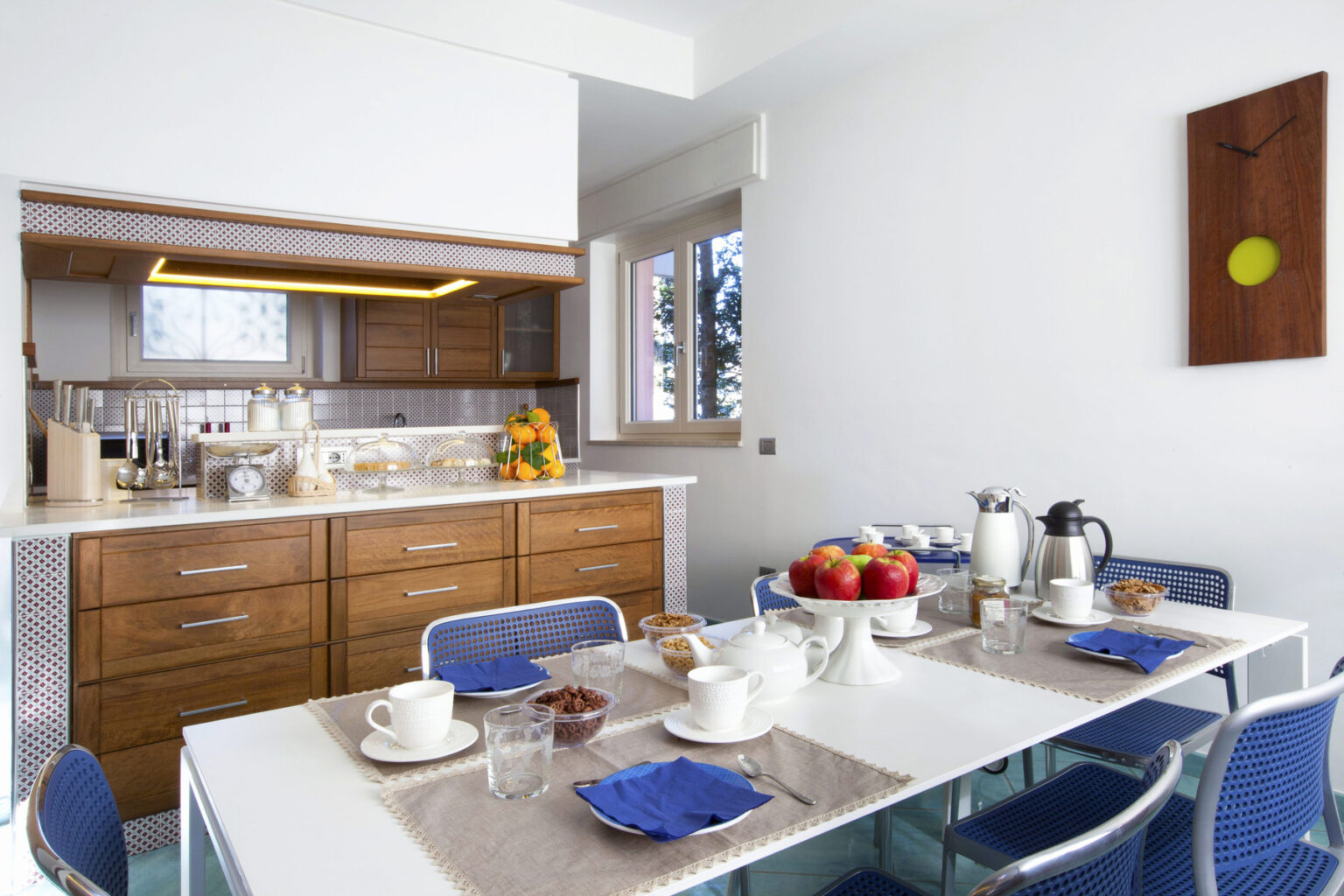 Modern kitchen and dining area featuring a sleek wooden countertop, blue dining chairs, and a table set with breakfast items like coffee, pastries, and fresh fruit. Bright natural light fills the space, enhancing the contemporary design. | Only on SopranoVillas