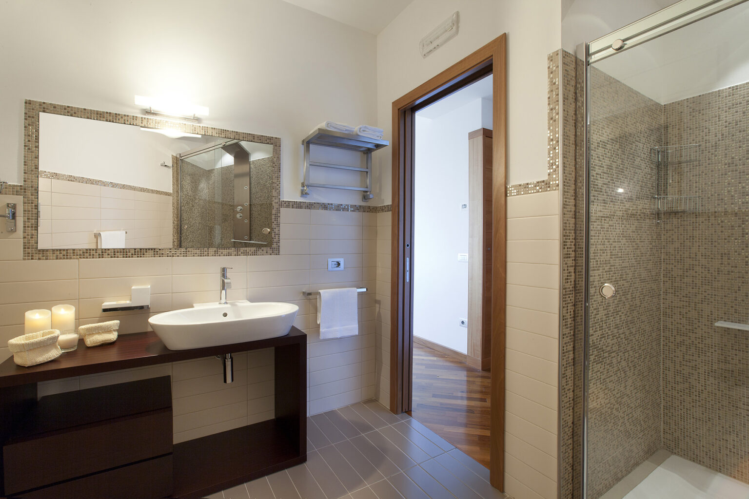 Modern bathroom featuring a sleek vanity with a sink, a large mirror, and a walk-in shower. The design includes neutral-toned tiles, wooden accents, and decorative candles for a stylish and relaxing atmosphere. | Only on SopranoVillas