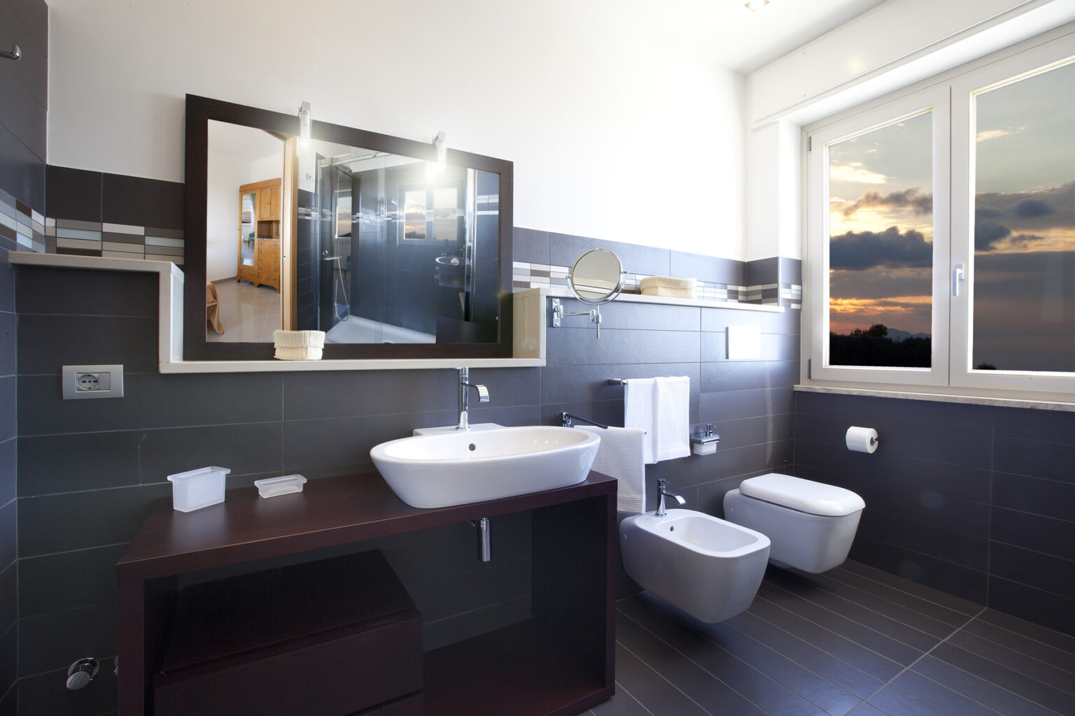 Modern bathroom interior featuring a sleek design, a large mirror, a stylish sink, and a bidet. Natural light streams in through a window, showcasing a scenic view at sunset. | Only on SopranoVillas