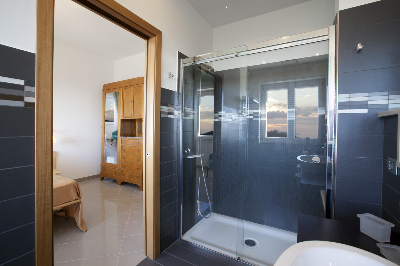 Modern bathroom with a glass shower enclosure, sleek gray tiles, and a view into a cozy bedroom featuring wooden furniture. | Only on SopranoVillas