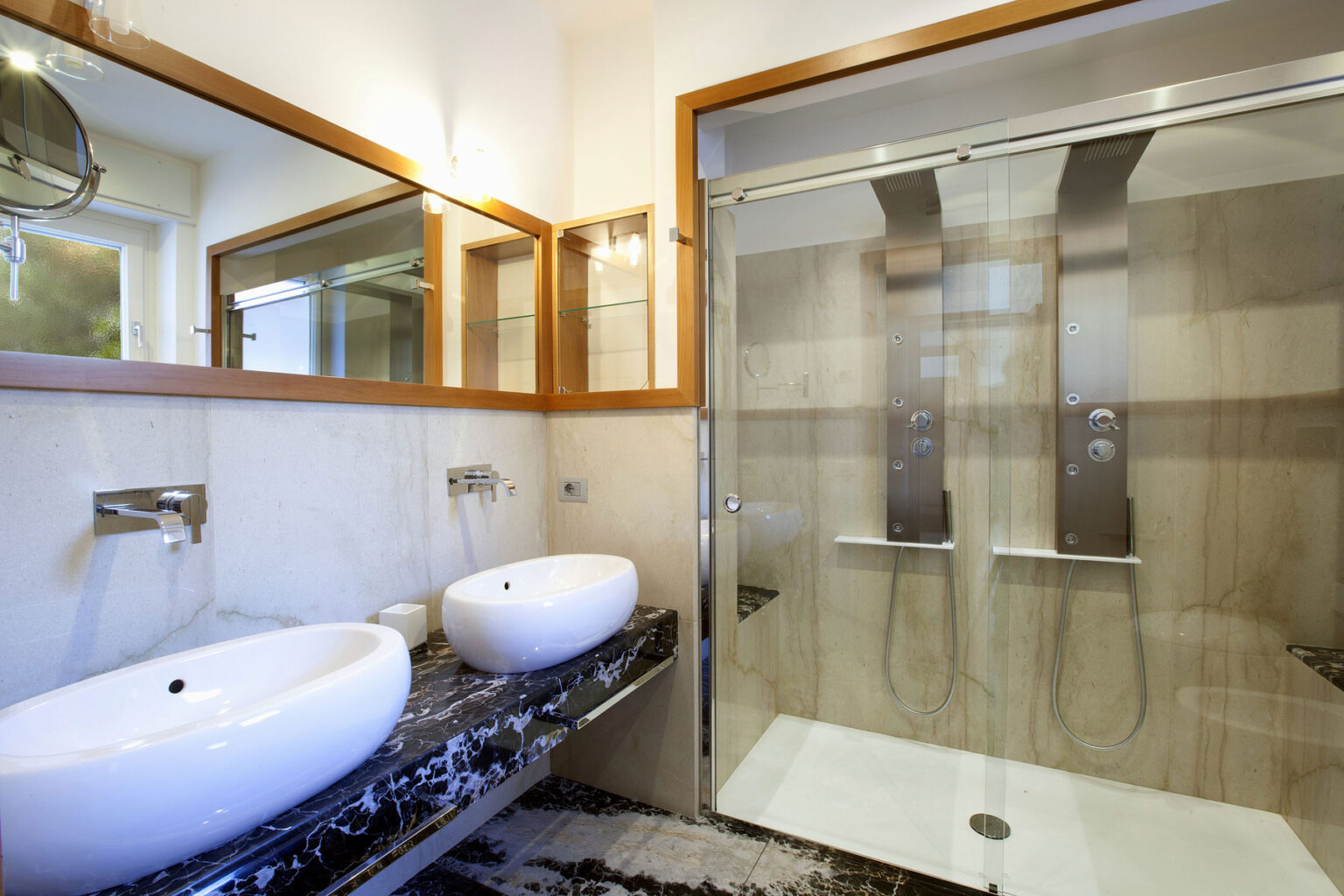 Modern bathroom featuring dual oval sinks on a sleek black marble countertop, contemporary fixtures, a spacious glass shower with dual shower panels, and stylish wooden accents. Ideal for luxury home design inspiration. | Only on SopranoVillas