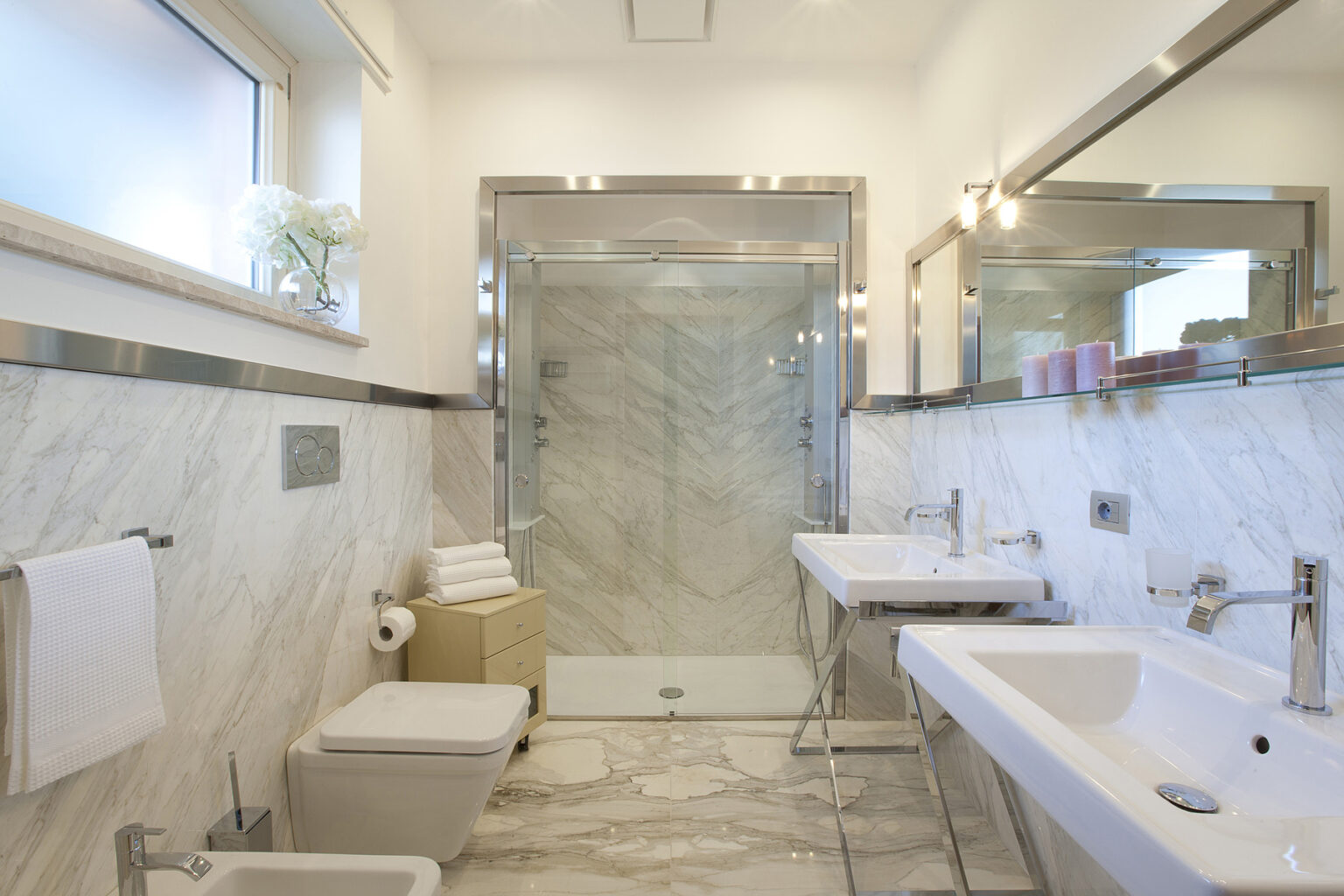 Modern bathroom featuring elegant marble walls, dual sinks, a glass shower enclosure, and stylish fixtures. Natural light floods the space through a window adorned with decorative flowers, enhancing the contemporary design. | Only on SopranoVillas