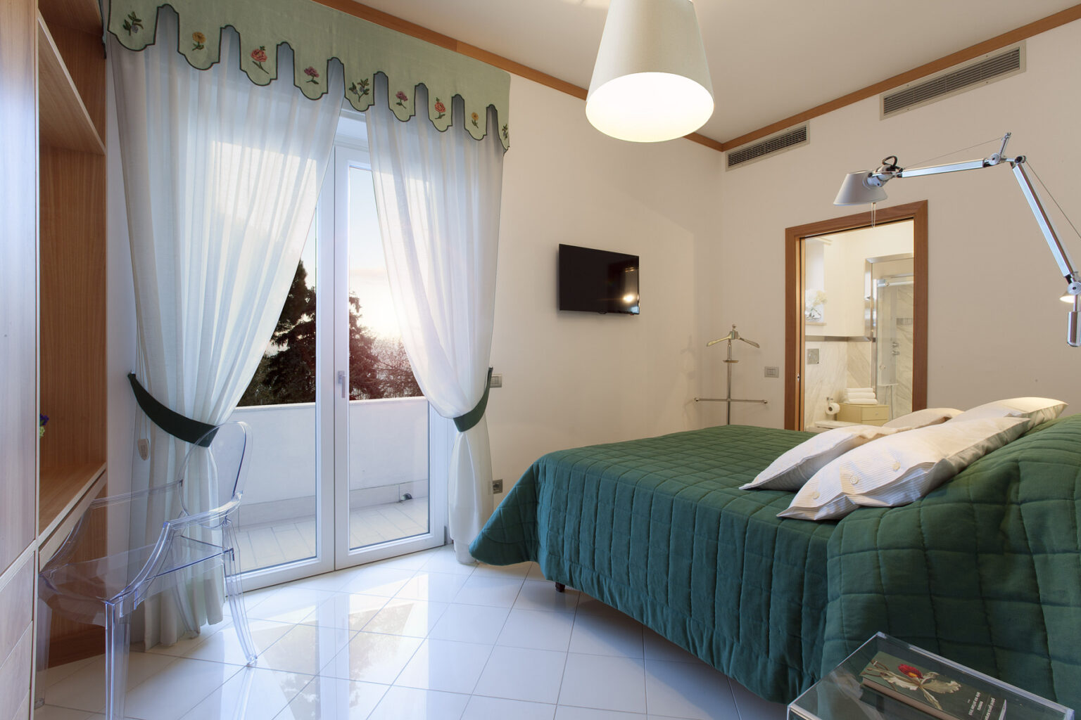 Bright and modern bedroom featuring a green quilted bedspread, large windows with sheer curtains, a flat-screen TV, and contemporary furniture including an acrylic chair and a coat stand. The room offers a view of the balcony and is designed with elegant decor elements. | Only on SopranoVillas