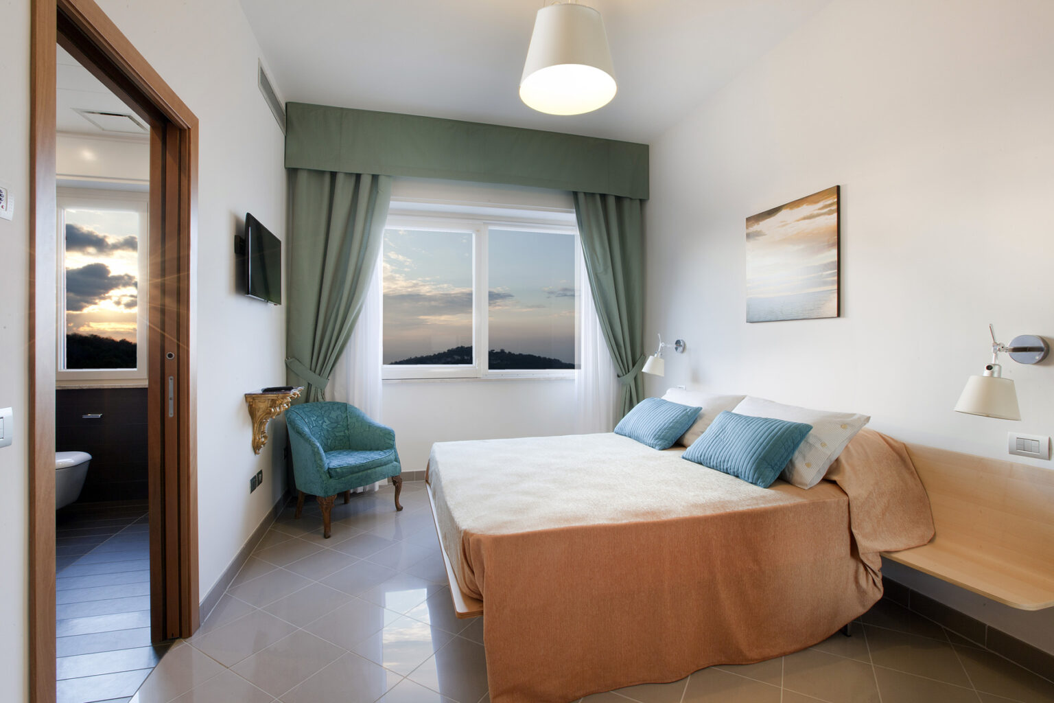 Modern hotel room featuring a queen-sized bed with decorative pillows, a cozy armchair, and large windows offering a scenic view. The room is adorned with soft lighting, elegant curtains, and contemporary decor, creating a relaxing atmosphere for guests. | Only on SopranoVillas