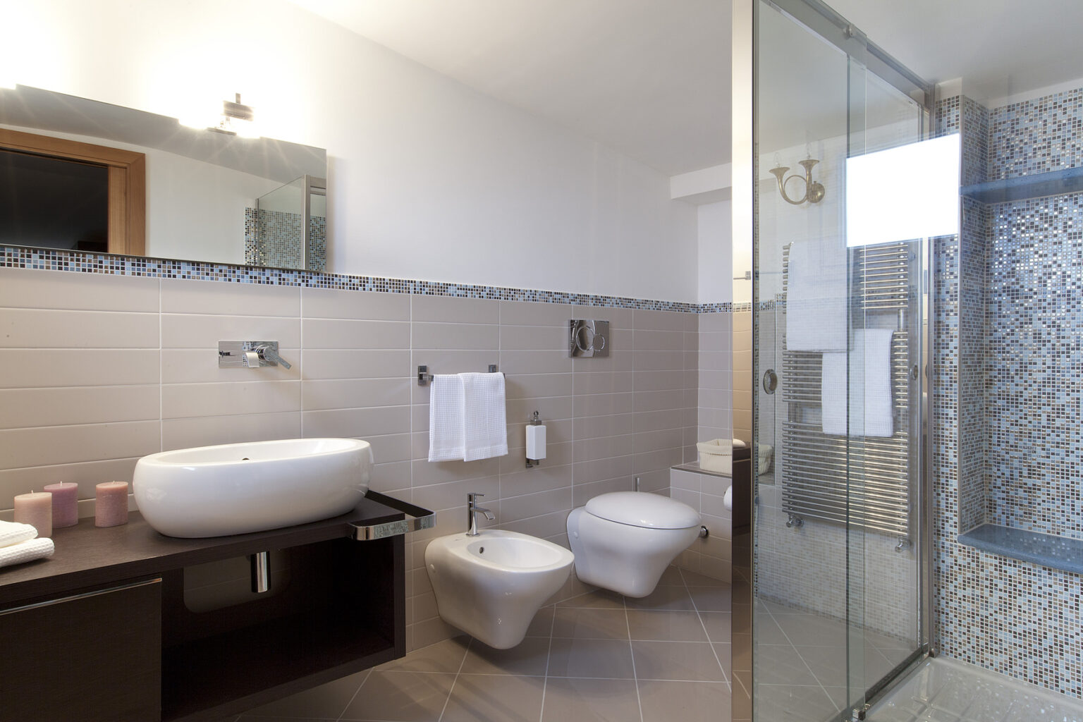 Modern bathroom interior featuring a sleek white sink, bidet, and toilet, complemented by a glass shower enclosure. The design includes elegant tiles with a mosaic accent and a stylish mirror, creating a contemporary and spacious atmosphere. | Only on SopranoVillas