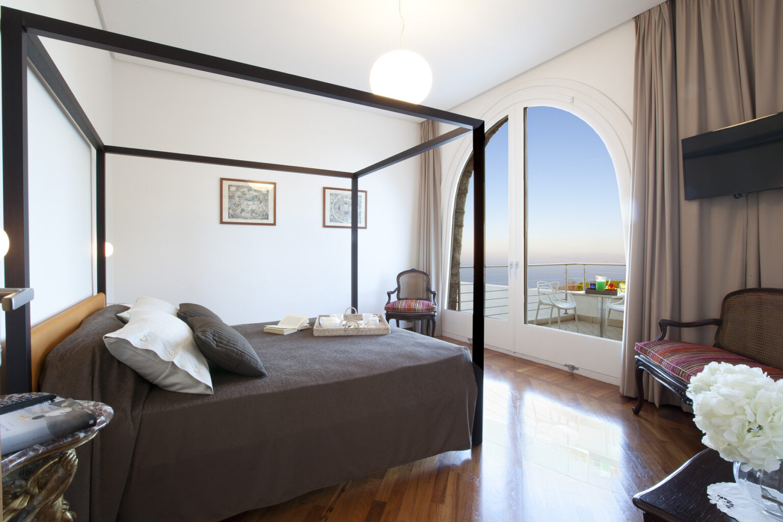 Elegant bedroom featuring a four-poster bed with dark bedding, stylish decor, and a large window offering a scenic view of the outdoors. The room includes vintage artwork and cozy seating, creating a serene atmosphere. | Only on SopranoVillas