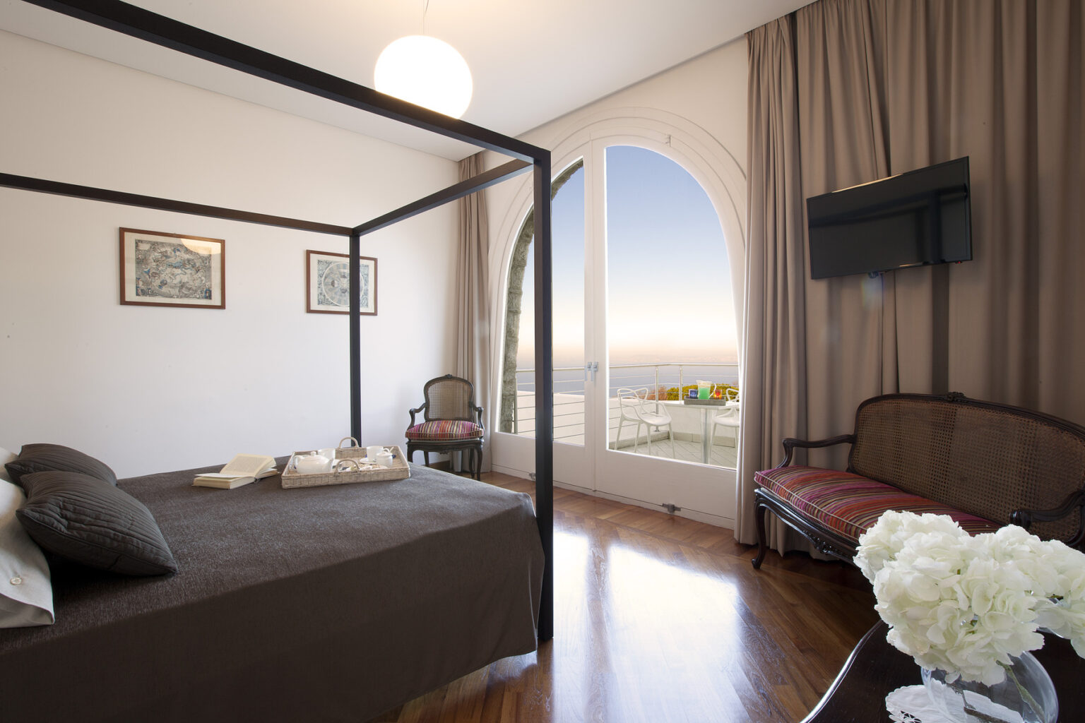 Elegant bedroom with a four-poster bed, hardwood floors, and large windows offering a scenic view. The room features modern decor, a flat-screen TV, and tasteful artwork, creating a cozy and inviting atmosphere. | Only on SopranoVillas