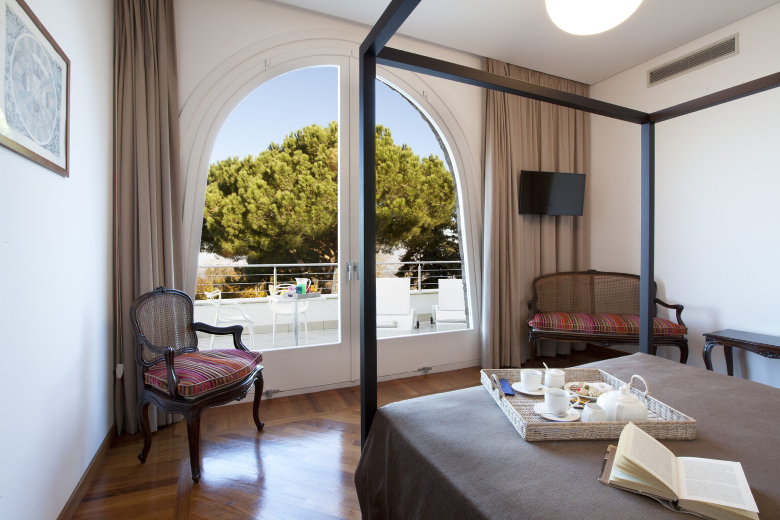 Elegant hotel room featuring a four-poster bed, antique seating, and large arched windows with a view of lush greenery. The room is designed with neutral tones and modern amenities, creating a cozy and inviting atmosphere. | Only on SopranoVillas