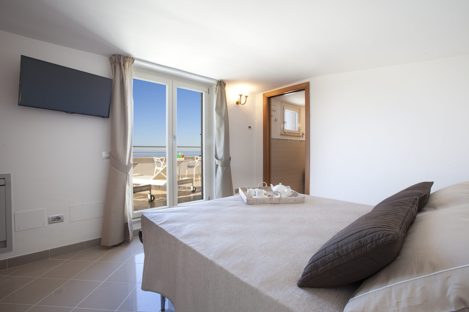 Cozy bedroom featuring a large bed with neutral bedding, a flat-screen TV mounted on the wall, and large windows offering a scenic view. The room is bright and airy, complemented by light-colored walls and elegant curtains. | Only on SopranoVillas