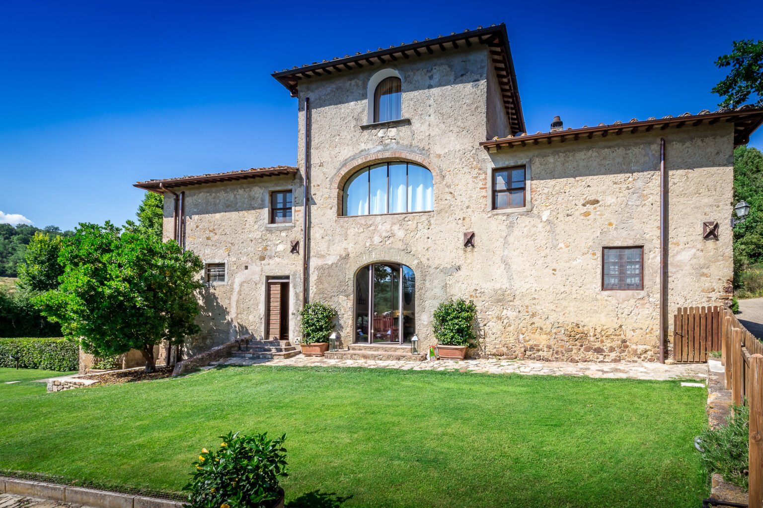 Beautiful rustic villa with stone facade and large arched windows, surrounded by a well-maintained lawn and greenery, set against a clear blue sky. Ideal for vacation rentals or countryside getaways. | Only on SopranoVillas
