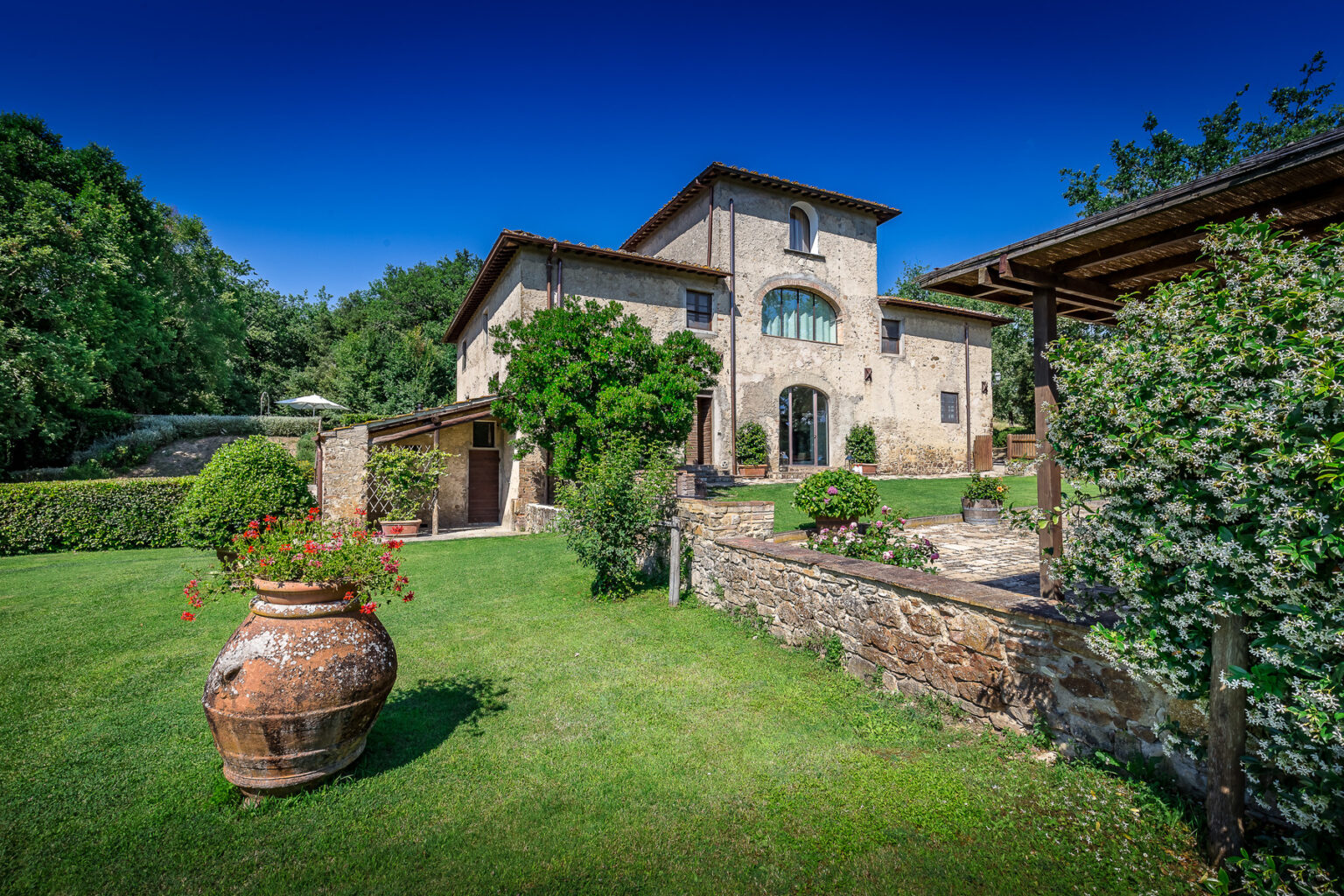 Rustic stone villa surrounded by lush greenery and gardens, featuring a large clay pot in the foreground and a clear blue sky above. Perfect for showcasing Italian countryside charm and outdoor living. | Only on SopranoVillas