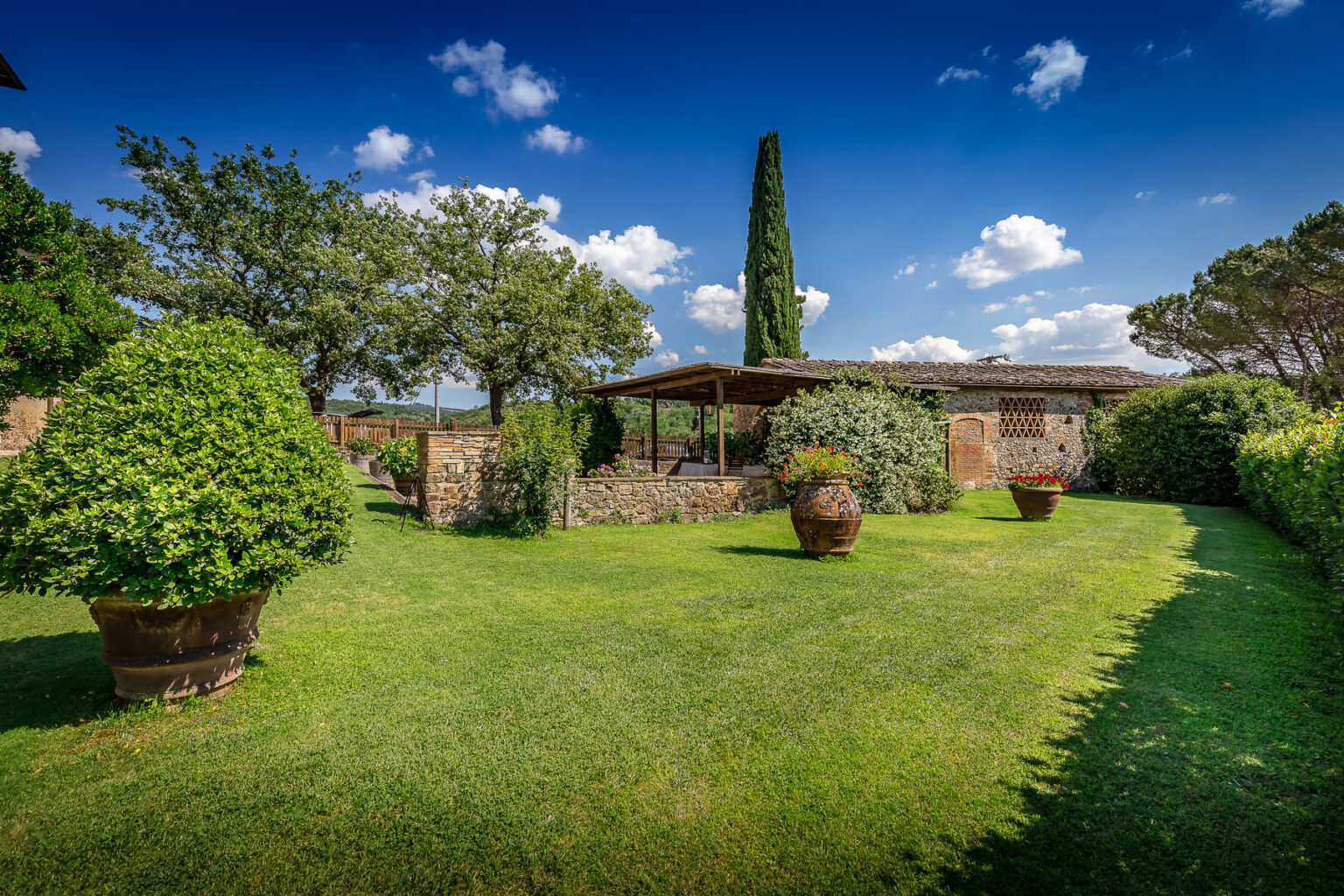 Scenic garden with lush green grass, decorative potted plants, and a stone building under a bright blue sky with fluffy clouds. Ideal for outdoor relaxation and gatherings. | Only on SopranoVillas