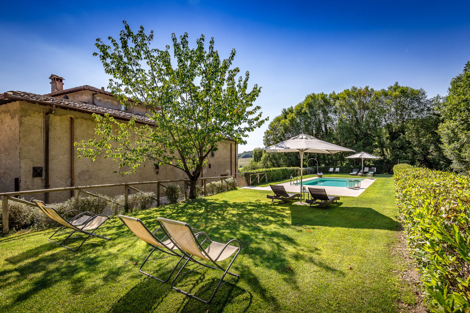 Scenic outdoor space featuring a well-maintained garden with a swimming pool, lounge chairs, and a shaded umbrella, set against a rustic building and lush greenery under a clear blue sky. Ideal for relaxation and leisure activities. | Only on SopranoVillas
