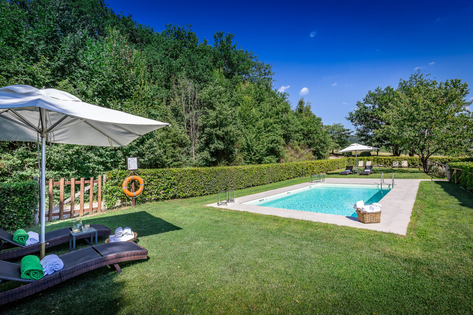 A serene outdoor pool area surrounded by lush greenery, featuring lounge chairs, umbrellas, and a clear blue sky, ideal for relaxation and leisure activities. | Only on SopranoVillas