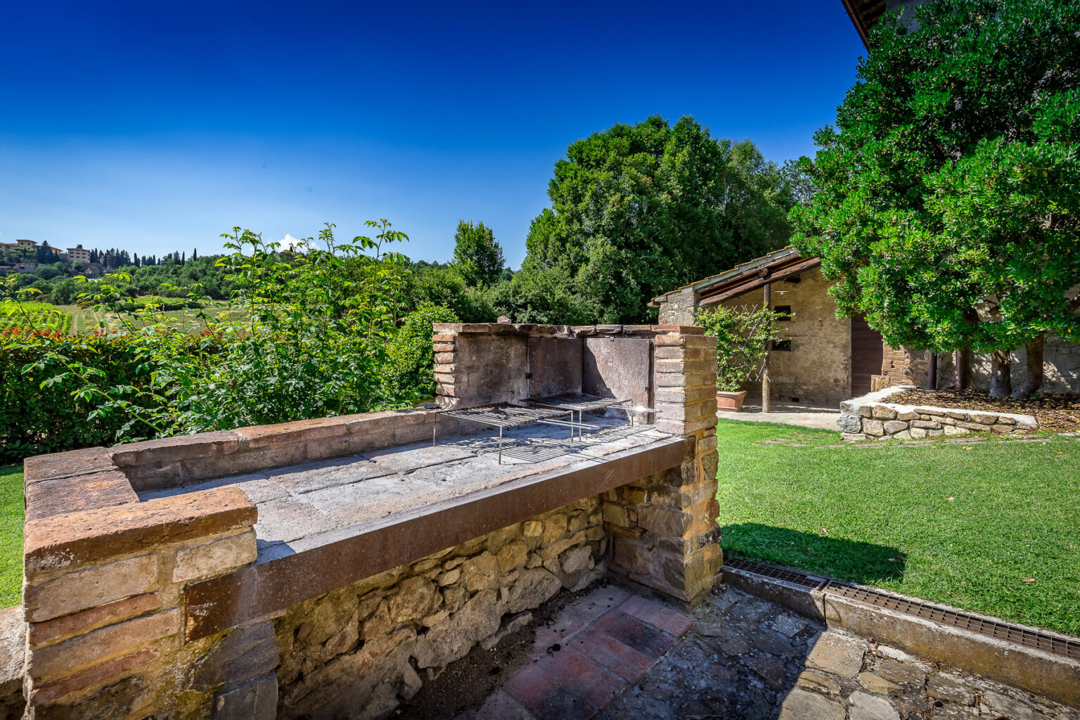 Outdoor barbecue area with a stone grill surrounded by lush greenery and a rustic building in the background, ideal for gatherings and outdoor cooking. | Only on SopranoVillas