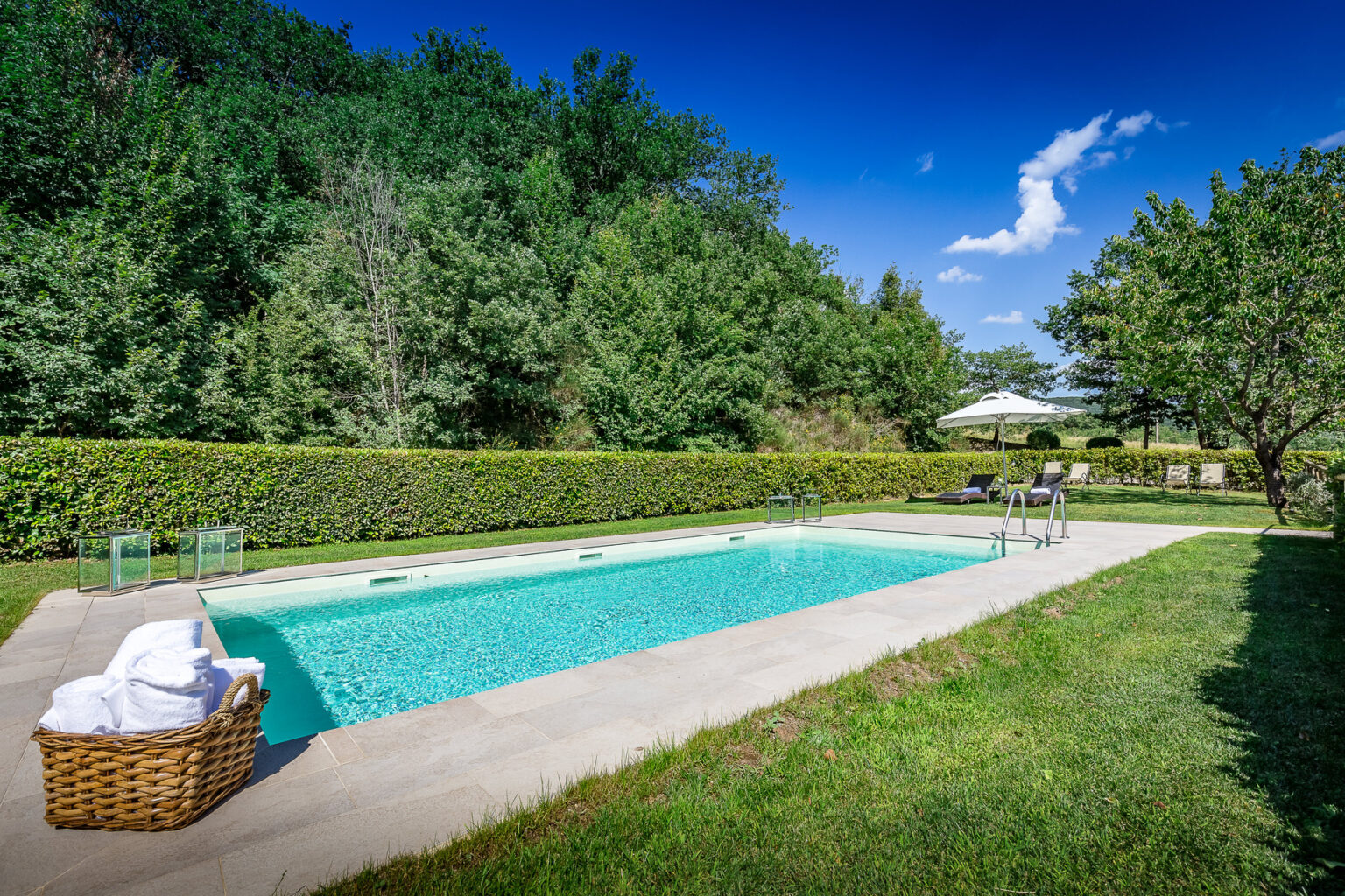 A serene outdoor swimming pool surrounded by lush greenery, featuring a basket of towels nearby and lounge chairs positioned for relaxation under a clear blue sky. | Only on SopranoVillas
