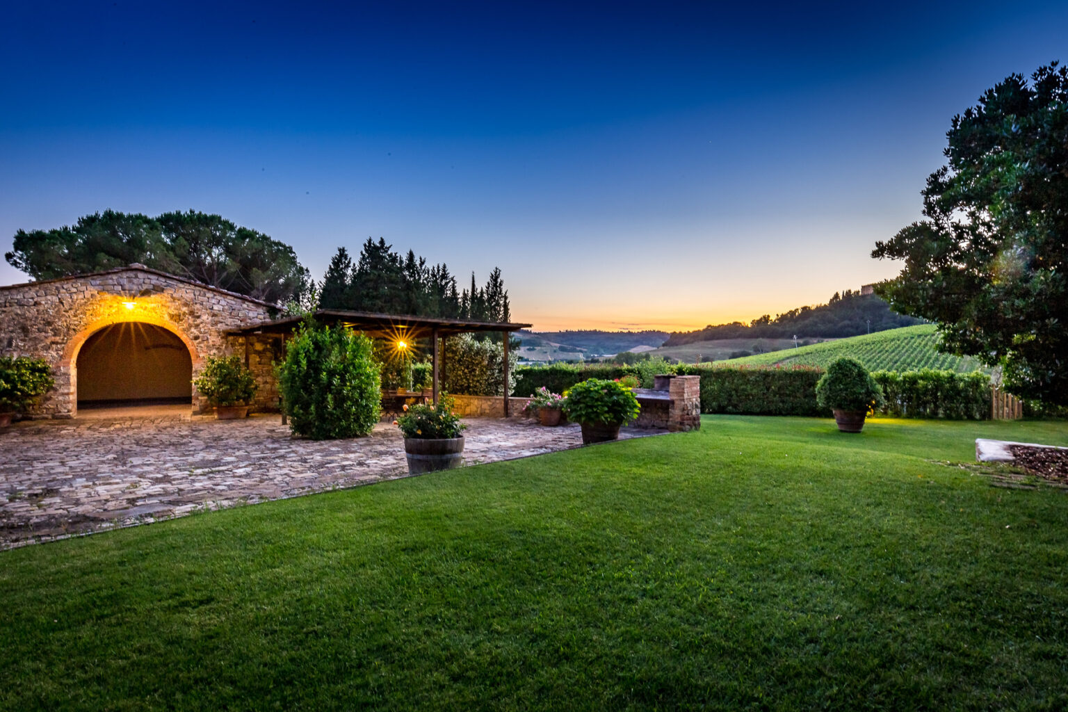 Scenic view of a rustic stone villa at sunset, surrounded by lush green lawns and vineyards, with a clear sky and trees in the background. Ideal for showcasing countryside retreats and wine country experiences. | Only on SopranoVillas