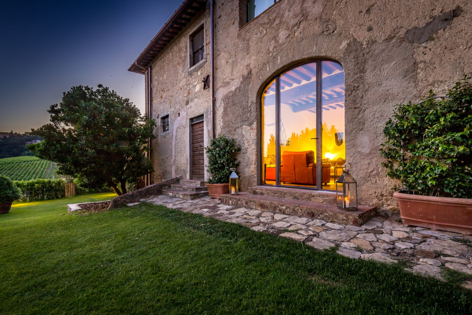 Rustic villa exterior at sunset, featuring a large arched window reflecting warm colors, surrounded by lush greenery and stone pathway. Ideal for showcasing beautiful countryside architecture and serene landscapes. | Only on SopranoVillas