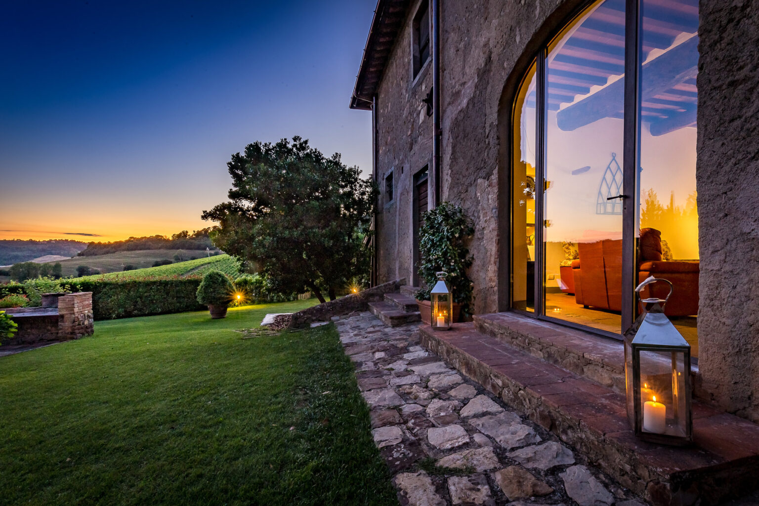 Scenic view of a rustic villa at sunset, featuring a stone pathway, lush green lawn, and warm glow from indoor lighting, surrounded by vineyards and trees. | Only on SopranoVillas