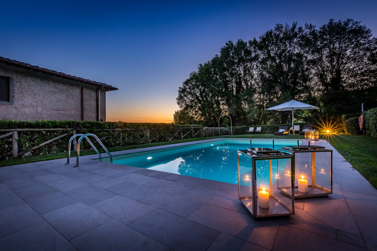 Serene evening view of a luxurious swimming pool surrounded by lush greenery, with elegant lanterns illuminating the area and a cozy seating area under an umbrella, capturing the tranquility of twilight. | Only on SopranoVillas
