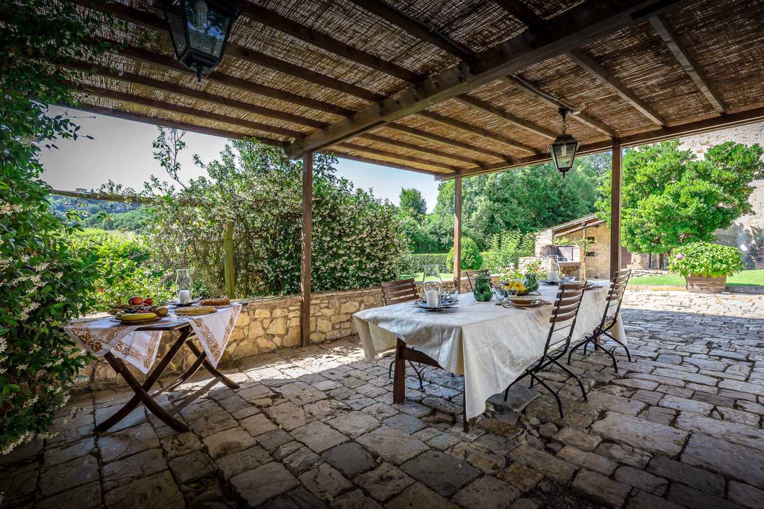 Outdoor dining area with a rustic stone floor, featuring a table set for a meal under a thatched roof, surrounded by lush greenery and a serene landscape. | Only on SopranoVillas