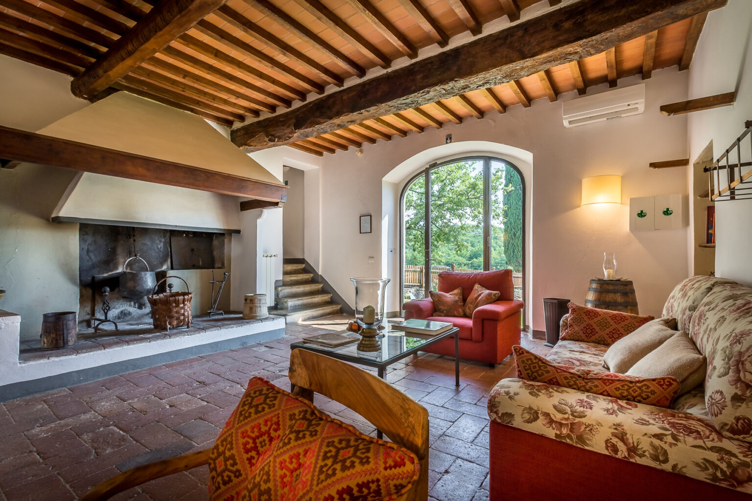 Spacious living room with rustic wooden beams, a cozy seating area featuring a floral sofa and red armchairs, a glass coffee table, and a traditional fireplace, all illuminated by natural light from large windows overlooking greenery. | Only on SopranoVillas