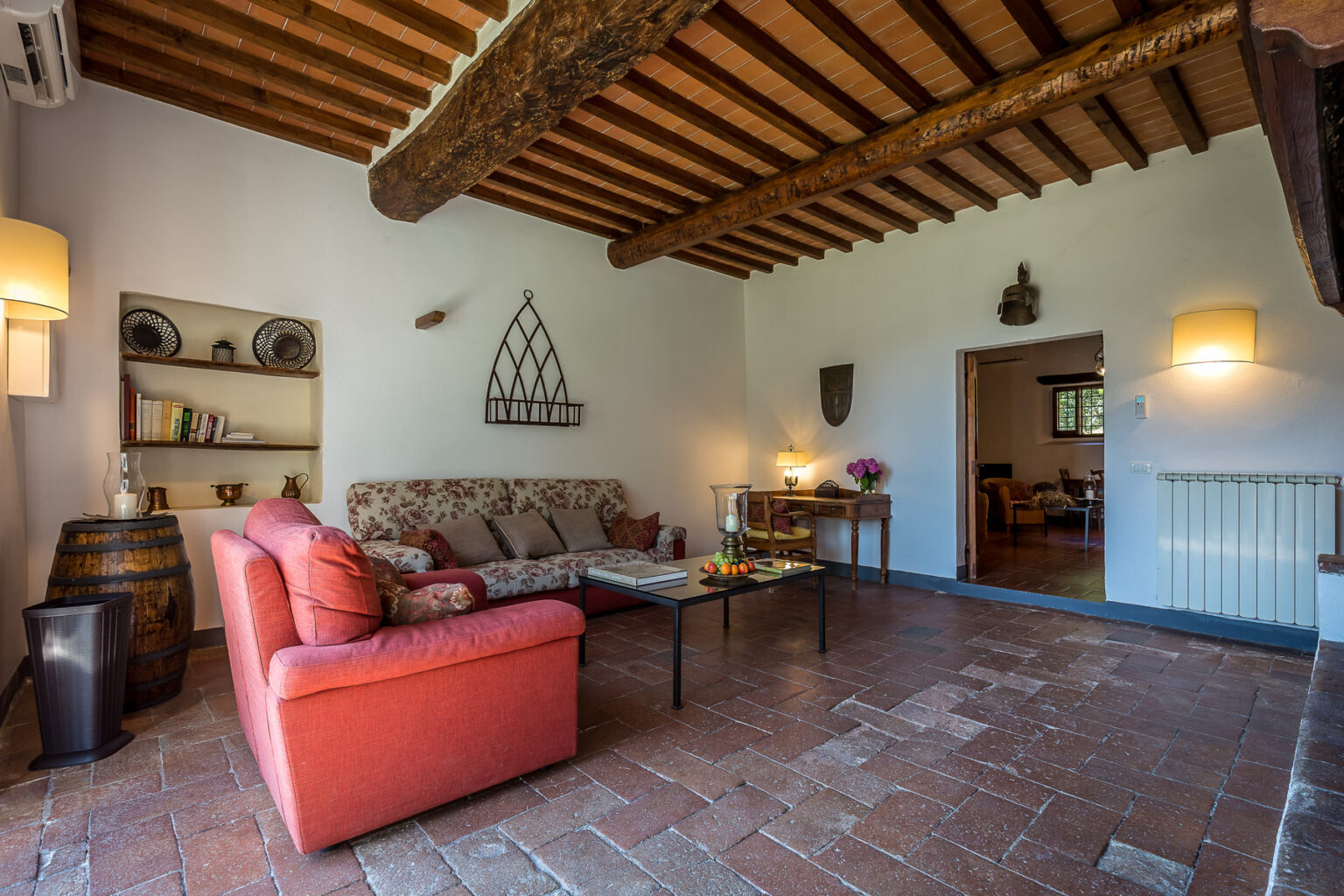 Cozy living room with rustic wooden beams, terracotta flooring, and a comfortable red sofa. Decorated with shelves, a decorative wall piece, and soft lighting, creating a warm and inviting atmosphere. Ideal for relaxation and gatherings. | Only on SopranoVillas