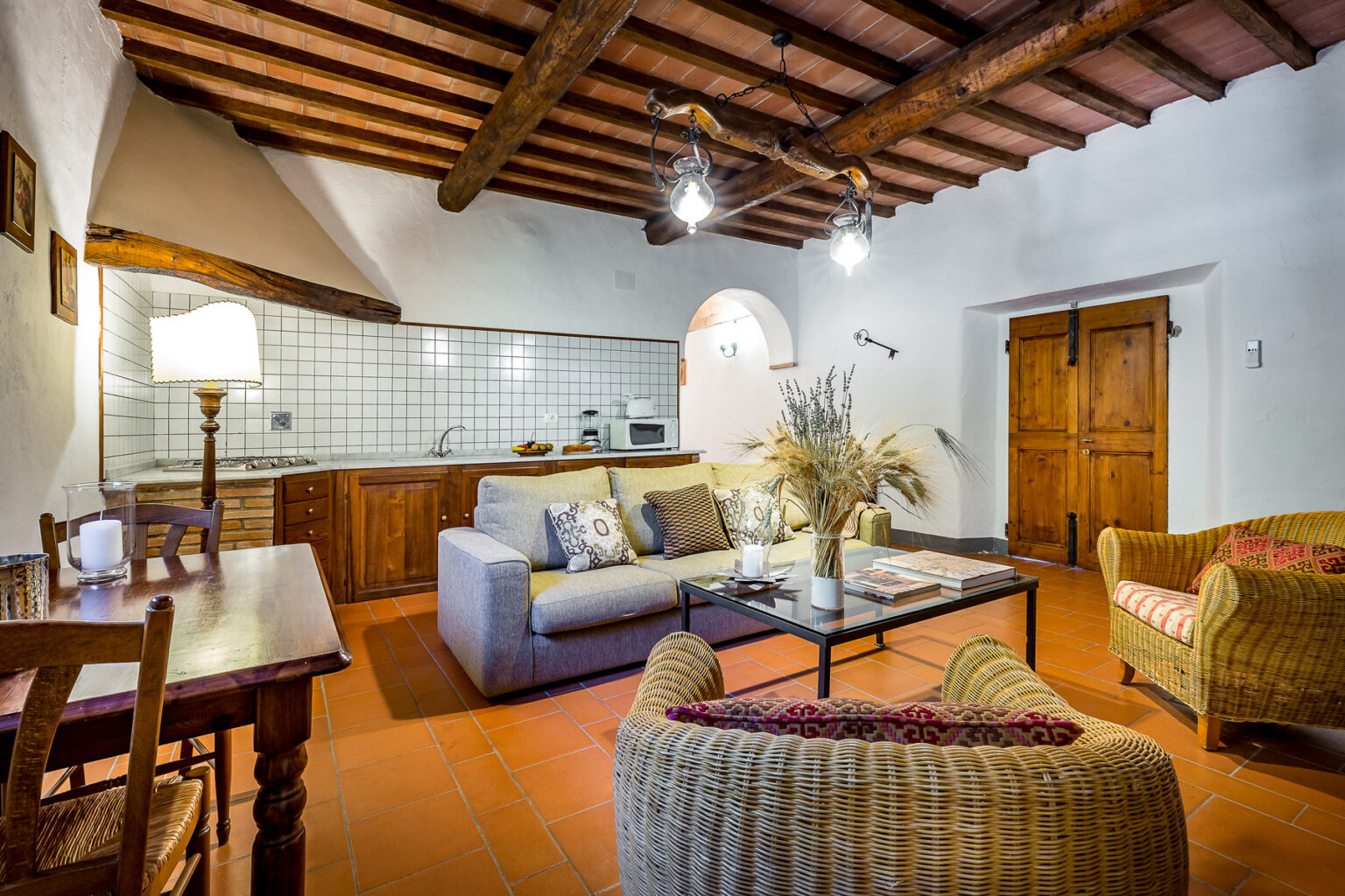 Cozy living room with rustic wooden beams, featuring a comfortable sofa, wicker chairs, and a wooden dining table. The space includes a modern kitchen area with tiled backsplash and warm lighting, creating an inviting atmosphere. | Only on SopranoVillas