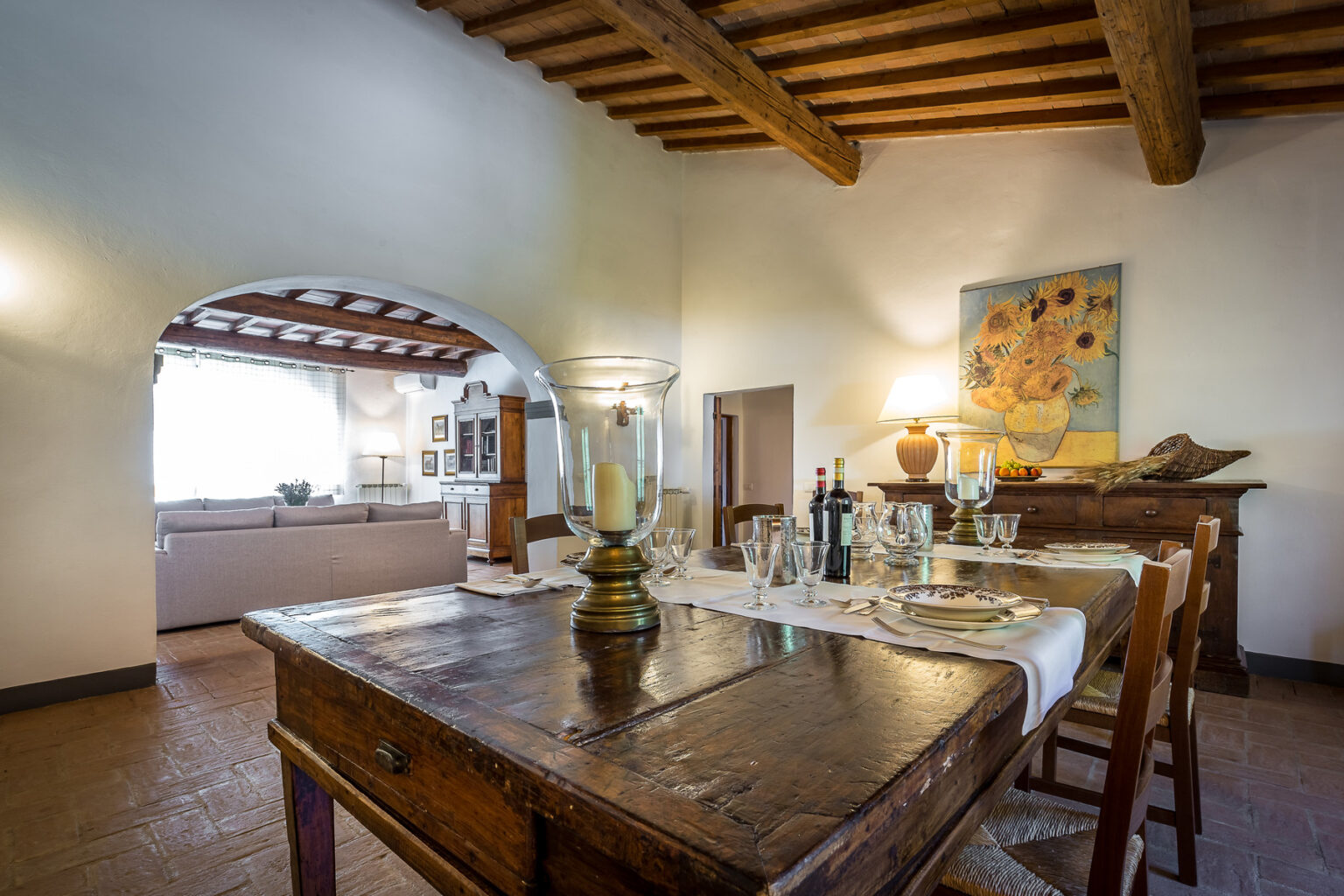 Rustic dining room with a wooden table set for a meal, featuring glassware and wine bottles, complemented by exposed wooden beams and a vibrant wall painting. The open layout connects to a spacious kitchen area. | Only on SopranoVillas