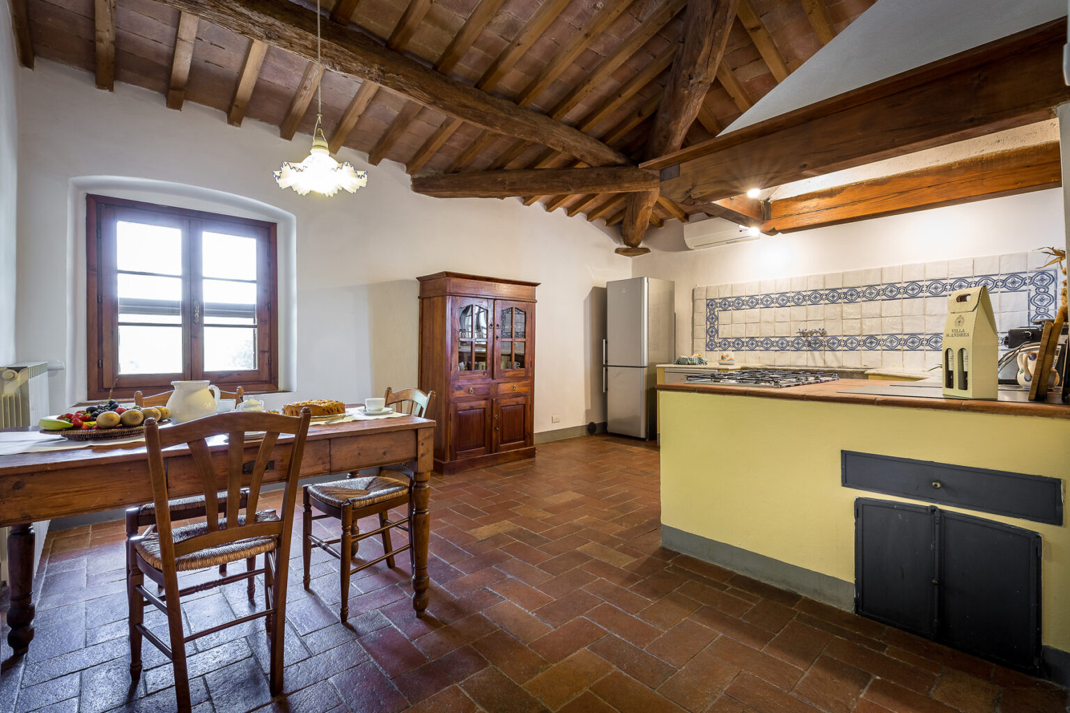 Spacious rustic kitchen featuring wooden beams, a dining table with chairs, and a modern kitchen area with appliances, complemented by natural light from large windows. | Only on SopranoVillas