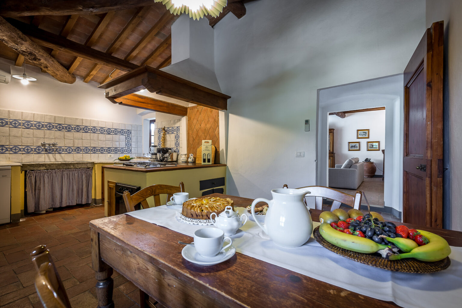 Rustic kitchen interior featuring a wooden dining table set with a teapot, cups, and a fruit platter, complemented by a cozy ambiance with exposed wooden beams and traditional decor. | Only on SopranoVillas