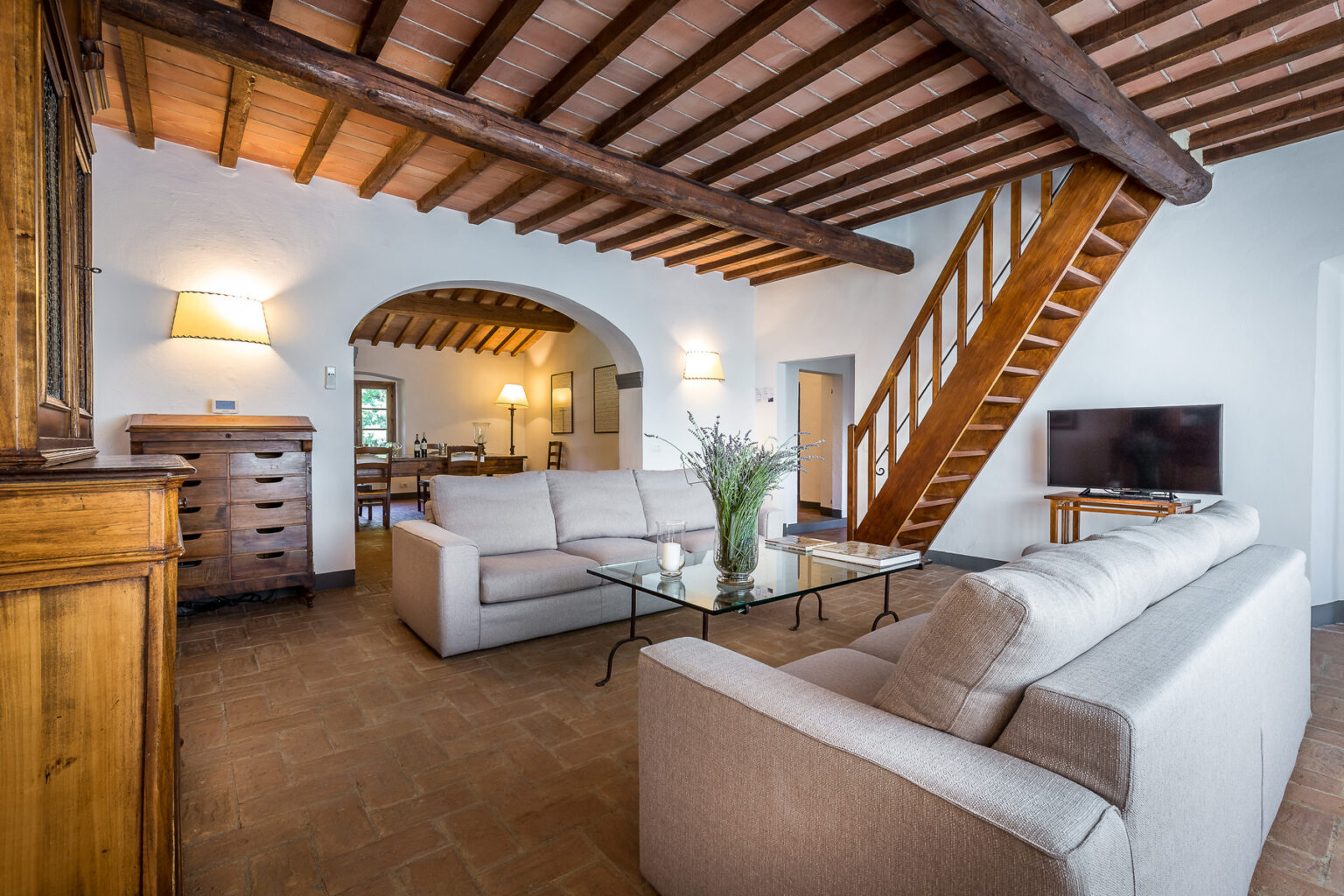 Spacious living room with wooden beams, modern furniture, and a staircase leading to an upper level, featuring a glass coffee table and a television, creating a cozy and inviting atmosphere. | Only on SopranoVillas