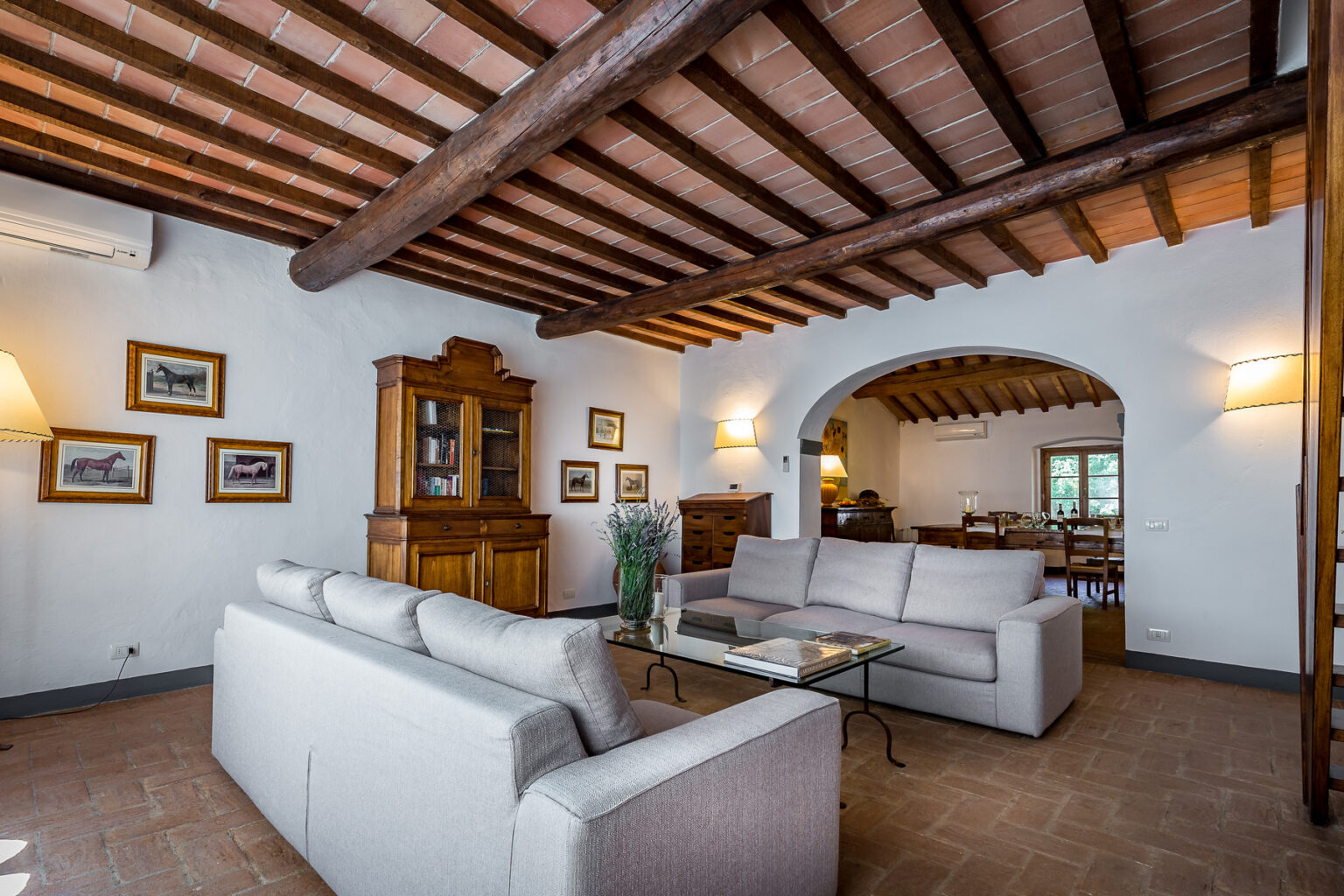 Cozy living room featuring a modern gray sofa, wooden furniture, and rustic ceiling beams. The space is adorned with framed artwork and includes a stylish coffee table. Natural light flows through an archway leading to a dining area, creating a warm and inviting atmosphere. Ideal for relaxation and family gatherings. | Only on SopranoVillas